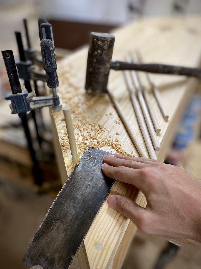 May include: A woodworking scene with a hand saw cutting a wooden dowel. A mallet, clamps, and several dowels are also visible on the wooden surface, along with wood shavings. The tools suggest a carpentry project in progress.