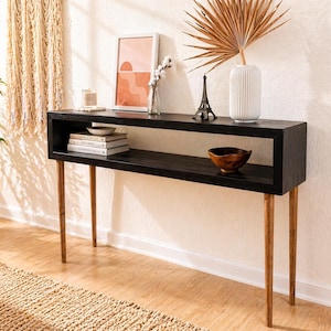May include: A black wooden console table with a rectangular top and a lower shelf. The table has four slender, light-colored wooden legs. Decorative items on the table include books, a small bowl, a framed print, and a vase with dried foliage.