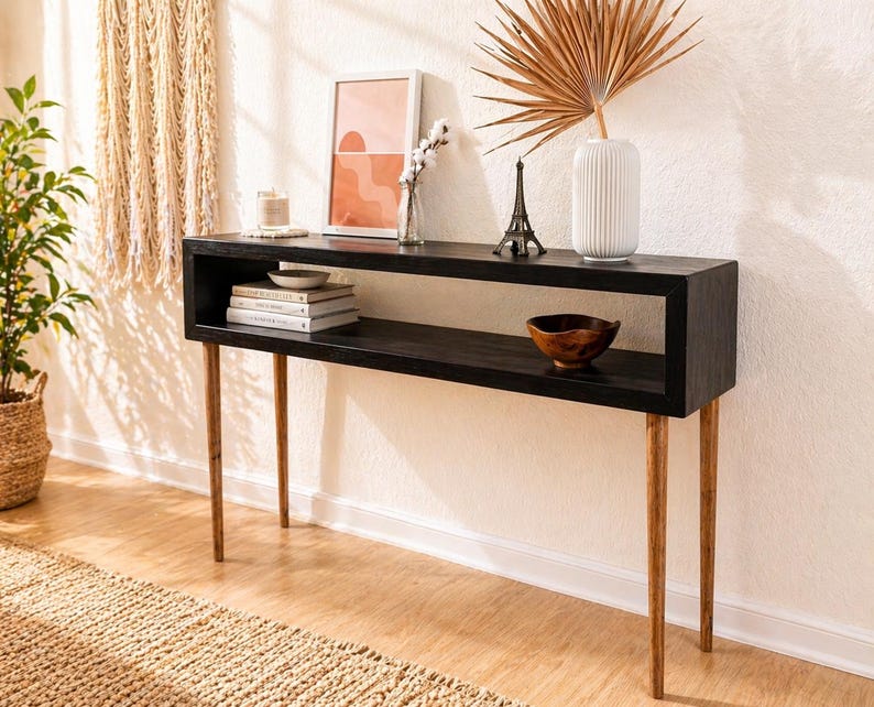 May include: A black wooden console table with a rectangular top and a lower shelf. The table has four slender, light-colored wooden legs. Decorative items on the table include books, a small bowl, a framed print, and a vase with dried foliage.