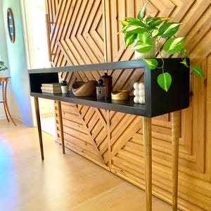 May include: A black console table with a rectangular top and four slender, angled wooden legs. The table has a shelf with decorative items, including books, a wooden bowl, and a small plant. A green and white plant sits on the right side of the table.