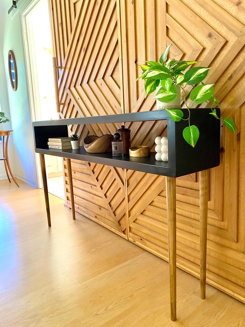 May include: A black console table with a rectangular top and four slender, angled wooden legs. The table has a shelf with decorative items, including books, a wooden bowl, and a small plant. A green and white plant sits on the right side of the table.