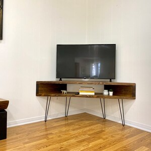 May include: A dark stained wooden TV stand with hairpin legs. The stand has an open shelf design with a rectangular shape. A flat-screen TV sits on top. The stand is in a corner of a room with hardwood floors.