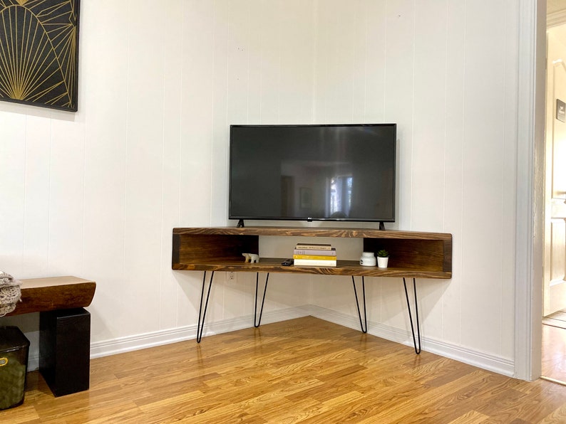 May include: A dark stained wooden TV stand with hairpin legs. The stand has an open shelf design with a rectangular shape. A flat-screen TV sits on top. The stand is in a corner of a room with hardwood floors.
