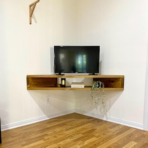 May include: A wooden floating corner shelf with a TV on top. The shelf is stained a warm brown and holds a small plant, books, and a candle. The shelf is mounted on a white wall with a wooden bracket above.
