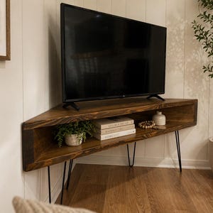 May include: A dark brown, corner TV stand with a black TV on top. The stand has a shelf with books, a small plant, and decorative items. The stand is supported by black hairpin legs. The room has a light wood floor and white walls.