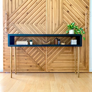 May include: A modern, black console table with a rectangular shelf and four slender, gold-toned legs. The shelf displays books, small plants, and decorative items. The backdrop is a wooden wall with a geometric pattern.