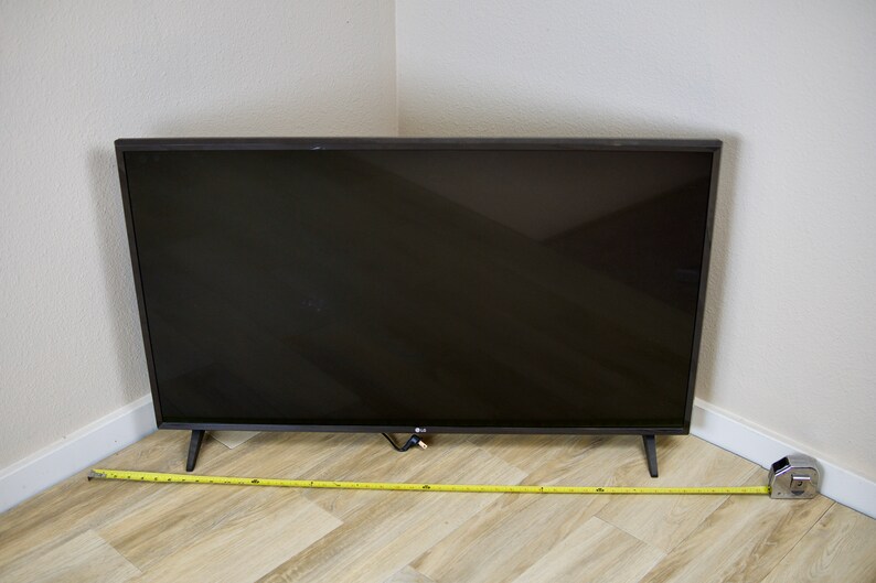 Puede incluir: Un televisor de pantalla plana LG negro con un marco delgado y cuatro patas negras. Una cinta m&eacute;trica amarilla se extiende por el suelo. El televisor est&aacute; colocado en una esquina contra paredes blancas.