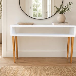 May include: A white rectangular console table with a hollow open shelf design and four tapered light brown wooden legs. The table surface holds a shallow wooden bowl and a book with the text Live Beautifully, alongside a small vase.
