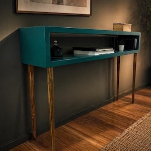 May include: Teal console table with a rectangular top and open shelf. The table has four slender, gold-tipped legs. A small black bottle, books, and a potted plant are visible on the shelf. The table is set against a dark gray wall.