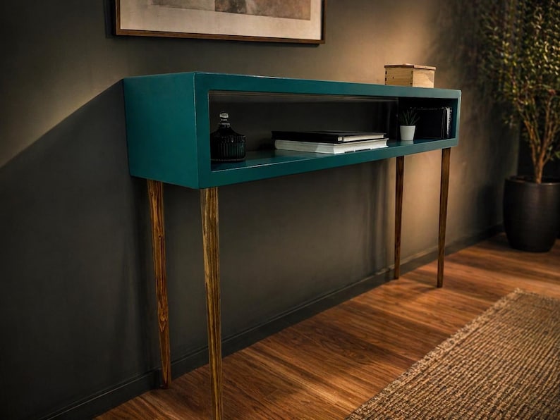 May include: Teal console table with a rectangular top and open shelf. The table has four slender, gold-tipped legs. A small black bottle, books, and a potted plant are visible on the shelf. The table is set against a dark gray wall.