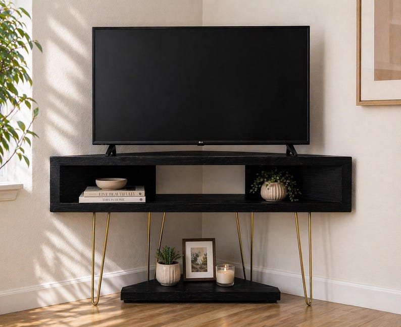 Puede incluir: Mueble de TV esquinero negro con un televisor de pantalla plana. El mueble tiene patas doradas de horquilla y un estante inferior con objetos decorativos, incluyendo una peque&ntilde;a planta, una foto enmarcada y una vela. El mueble es de madera.