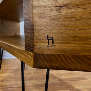 May include: A handcrafted wooden coffee table with a natural wood grain finish. The table features a rectangular design with a lower shelf and is supported by black hairpin legs. The table has a dark logo on the side.