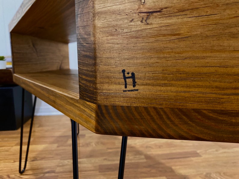 May include: A handcrafted wooden coffee table with a natural wood grain finish. The table features a rectangular design with a lower shelf and is supported by black hairpin legs. The table has a dark logo on the side.