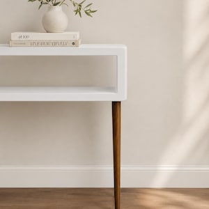 May include: A white modern side table with a rectangular top and a lower shelf. The table has four angled, brown wooden legs. A small white vase with greenery and two books are on top of the table.