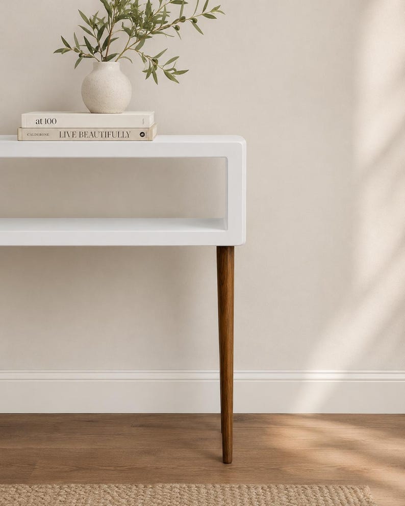 May include: A white modern side table with a rectangular top and a lower shelf. The table has four angled, brown wooden legs. A small white vase with greenery and two books are on top of the table.
