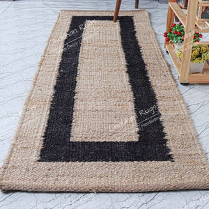 May include: A natural fiber rug with a black border. The rug is made of jute and has a handwoven texture. The rug is rectangular and has a simple design.