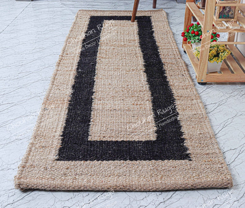 May include: A natural fiber rug with a black border. The rug is made of jute and has a handwoven texture. The rug is rectangular and has a simple design.