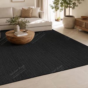 May include: A large, rectangular area rug featuring a solid, deep black color and a textured, woven surface. The rug is positioned flat on a light-colored floor, extending from the foreground toward the center of the room under a wooden coffee table.