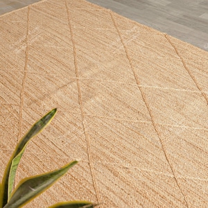 Puede incluir: Una alfombra de fibra natural con un patrón de diamantes tejido en la superficie. La alfombra es de color marrón claro y tiene un aspecto natural y rústico.