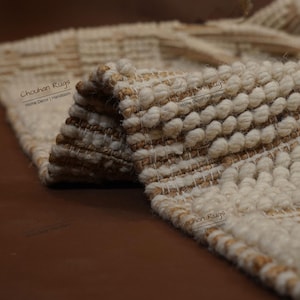 Puede incluir: Primer plano de una alfombra beige y marrón texturizada. La alfombra presenta un diseño tejido con elementos redondeados en relieve y patrones lineales. La alfombra está doblada, revelando su construcción en capas. La alfombra es adecuada para la decoración del hogar.