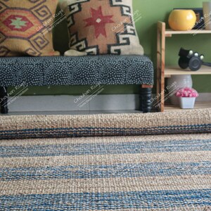 K&ouml;nnte beinhalten: Ein zusammengerollter Naturfaser-Teppich mit blauen Streifen. Der Teppich liegt vor einer Bank mit zwei gemusterten Kissen und einem Holzregal mit einer gelben Schale und einem schwarzen Gegenstand.