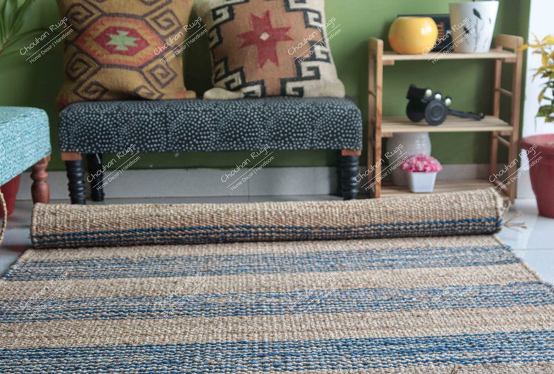 K&ouml;nnte beinhalten: Ein zusammengerollter Naturfaser-Teppich mit blauen Streifen. Der Teppich liegt vor einer Bank mit zwei gemusterten Kissen und einem Holzregal mit einer gelben Schale und einem schwarzen Gegenstand.