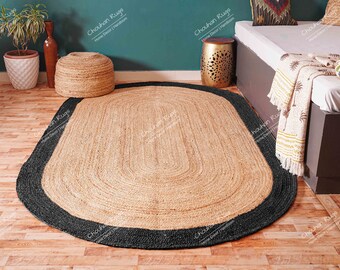 May include: Oval jute rug with a black border. The rug has a natural tan center and a dark border. The rug is made of woven jute fibers. The rug is on a wooden floor.