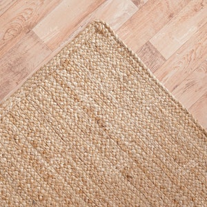 May include: A close-up of a natural fiber rug made from woven jute. The rug is a light beige color and has a rough, textured surface.
