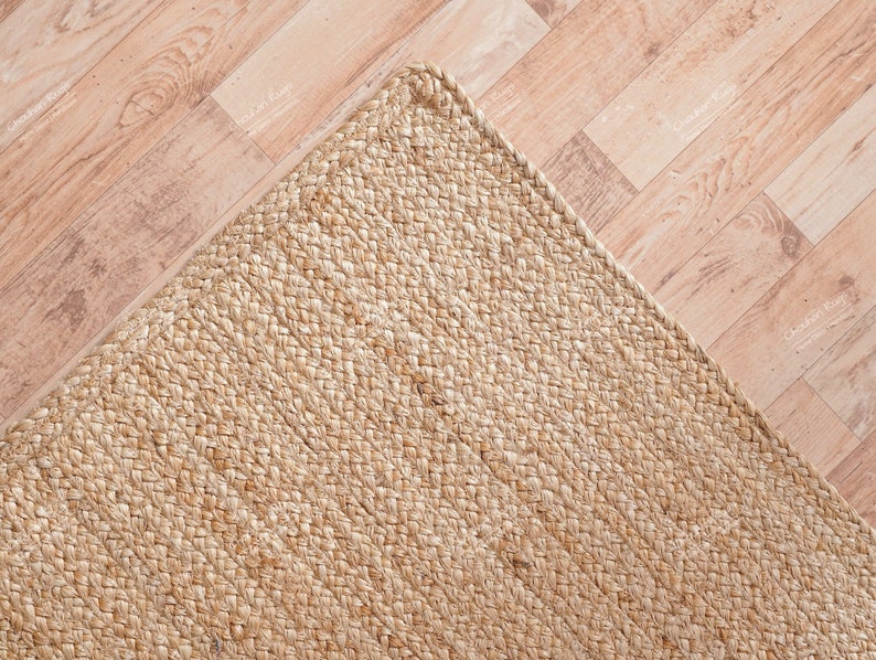May include: A close-up of a natural fiber rug made from woven jute. The rug is a light beige color and has a rough, textured surface.