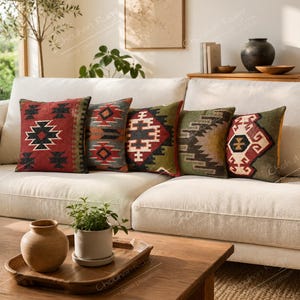 May include: Five square throw pillows arranged in a row on a cream-colored sofa. Each pillow features a distinct woven geometric pattern in various color combinations including red, olive green, charcoal, and cream, showcasing intricate textile designs.