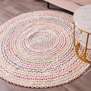 Puede incluir: Una alfombra redonda trenzada con un patrón en espiral de tela color blanco hueso entrelazada con pequeñas motas de colores rojo, amarillo, azul, verde y morado. La alfombra tiene una construcción gruesa y texturizada con un tejido circular.