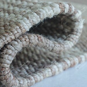 K&ouml;nnte beinhalten: Nahaufnahme eines Naturfaser-Teppichs mit gewebter Struktur. Der Teppich ist aus Jute und hat eine hellbraune Farbe. Der Teppich ist in der Mitte gefaltet, wodurch die Innenseite des Gewebes sichtbar wird.