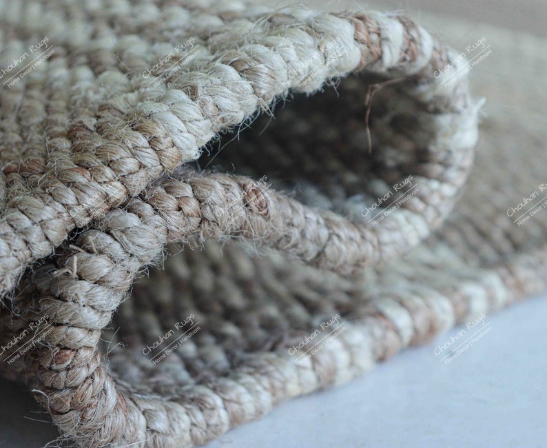 K&ouml;nnte beinhalten: Nahaufnahme eines Naturfaser-Teppichs mit gewebter Struktur. Der Teppich ist aus Jute und hat eine hellbraune Farbe. Der Teppich ist in der Mitte gefaltet, wodurch die Innenseite des Gewebes sichtbar wird.
