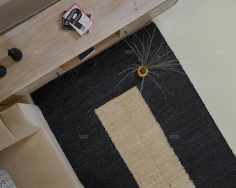 May include: Overhead view of a black and beige rectangular rug. The rug has a black border and a beige rectangular center. A small plant in a yellow pot sits on the rug. A light-colored wooden table is in the upper left corner.