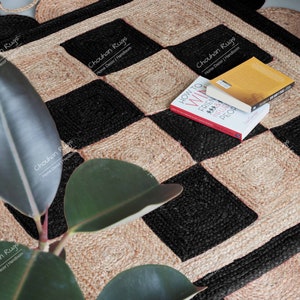 Könnte beinhalten: Ein gewebter Jute-Teppich mit einem Schachbrettmuster aus schwarzen und naturfarbenen beigen Quadraten. Der Teppich hat einen gewellten Rand und ist mit zwei Büchern verziert. Der Text "Chouhan Rugs" ist sichtbar.