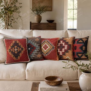May include: Five square throw pillows arranged on a cream sofa, each featuring distinct geometric patterns in red, blue, brown, and black. The designs include diamond and cross motifs, showcasing a woven texture across the set of decorative cushions.