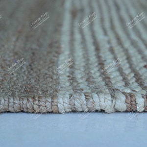 K&ouml;nnte beinhalten: Nahaufnahme eines Naturfaser-Teppichs mit einem gestreiften Muster. Der Teppich ist aus Jute und hat eine hellbraune und wei&szlig;e Farbgebung. Der Teppich ist mit einer dichten Webart gewebt und hat eine leicht strukturierte Oberfl&auml;che.