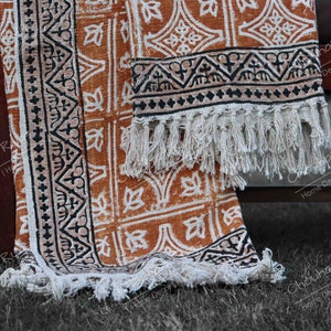 May include: A handwoven cotton throw blanket with a geometric pattern in shades of orange, black, and white. The blanket has a fringe border and is folded on a wooden surface.