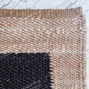 May include: Close-up of a woven jute rug with a black and beige geometric pattern. The rug has a natural, earthy feel and is perfect for adding a touch of texture to any room.