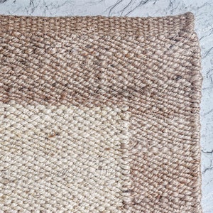 May include: A close-up of a woven rug with a diagonal pattern. The rug is made of natural fibers and has a light brown and cream color.