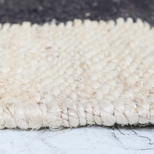 Op de afbeelding: Een close-up van een natuurlijk vezel tapijt gemaakt van geweven jute. Het tapijt is licht beige van kleur en heeft een ruw, getextureerd oppervlak.