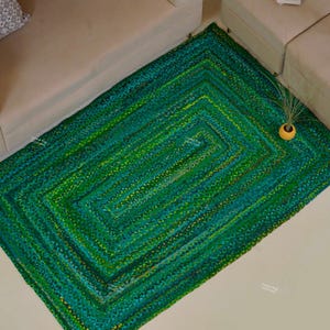 Könnte beinhalten: Ein rechteckiger grüner Teppich mit geflochtenem Design. Der Teppich weist Grün- und Gelbtöne auf und erzeugt ein strukturiertes Aussehen. Der Teppich liegt auf einem hellen Boden, in der Nähe eines beigen Sofas und einer passenden Ottomane.