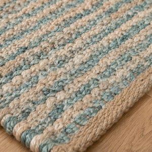 Peut inclure: Un tapis tissé rectangulaire présentant des rayures alternées de fibres bleu clair et crème. La texture est épaisse et tressée, avec une bordure beige unie le long du bord du tapis, mettant en valeur une construction dense et nouée.