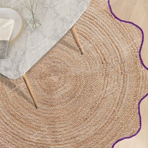 May include: A round, woven jute rug with a scalloped purple border. The rug is tan and has a spiral design in the center. A marble-topped table with wooden legs is partially visible in the upper left corner.