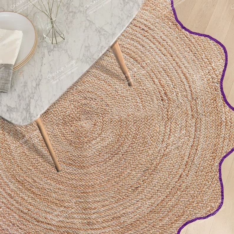 May include: A round, woven jute rug with a scalloped purple border. The rug is tan and has a spiral design in the center. A marble-topped table with wooden legs is partially visible in the upper left corner.