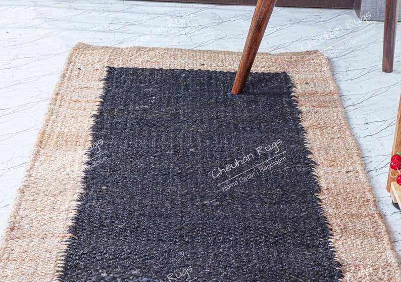 May include: A black and natural jute rug with a black center and a natural jute border. The rug has a rough, textured surface. The text "Chouchan Rugs" is printed in white on the black center of the rug.