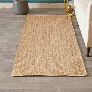 "Captured from a top-down view on a white background, this durable mat showcases the inherent beauty and linear texture of natural jute fiber. The consistent, dense braiding along the length ensures resilience and provides a soft, earthy aesthetic ideal for layering or high-traffic areas."