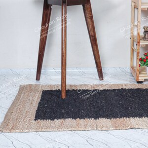 May include: A black and beige jute rug with a black center section. The rug is placed on a white tiled floor. A wooden stool with a brown top is standing on the rug.