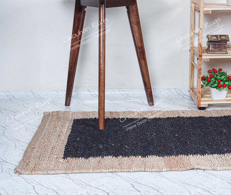 May include: A black and beige jute rug with a black center section. The rug is placed on a white tiled floor. A wooden stool with a brown top is standing on the rug.