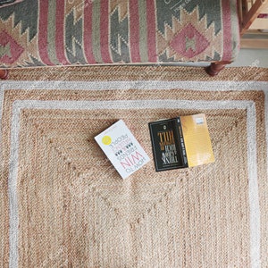 May include: A rectangular jute rug with a natural beige color and a white border. Two books are placed on the rug. The rug is surrounded by a patterned bench, a plant in a terracotta pot, and a wooden frame.